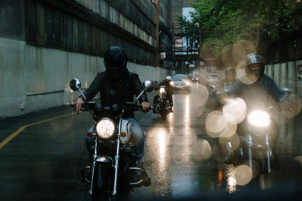Motorrijders rijden door een stedelijke omgeving bij nat wegdek en schemering.